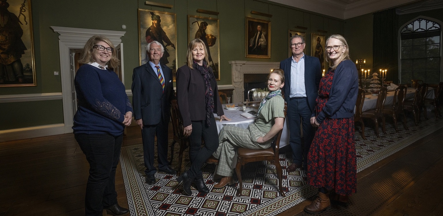 Pictured are representatives from Visit County Durham, Durham County Council and other Memorable Moments partners in the Long Dining Room in Auckland Castle. They are stood and seated in from of a table, which is set for dinner. There are large paintings on the wall behind them.