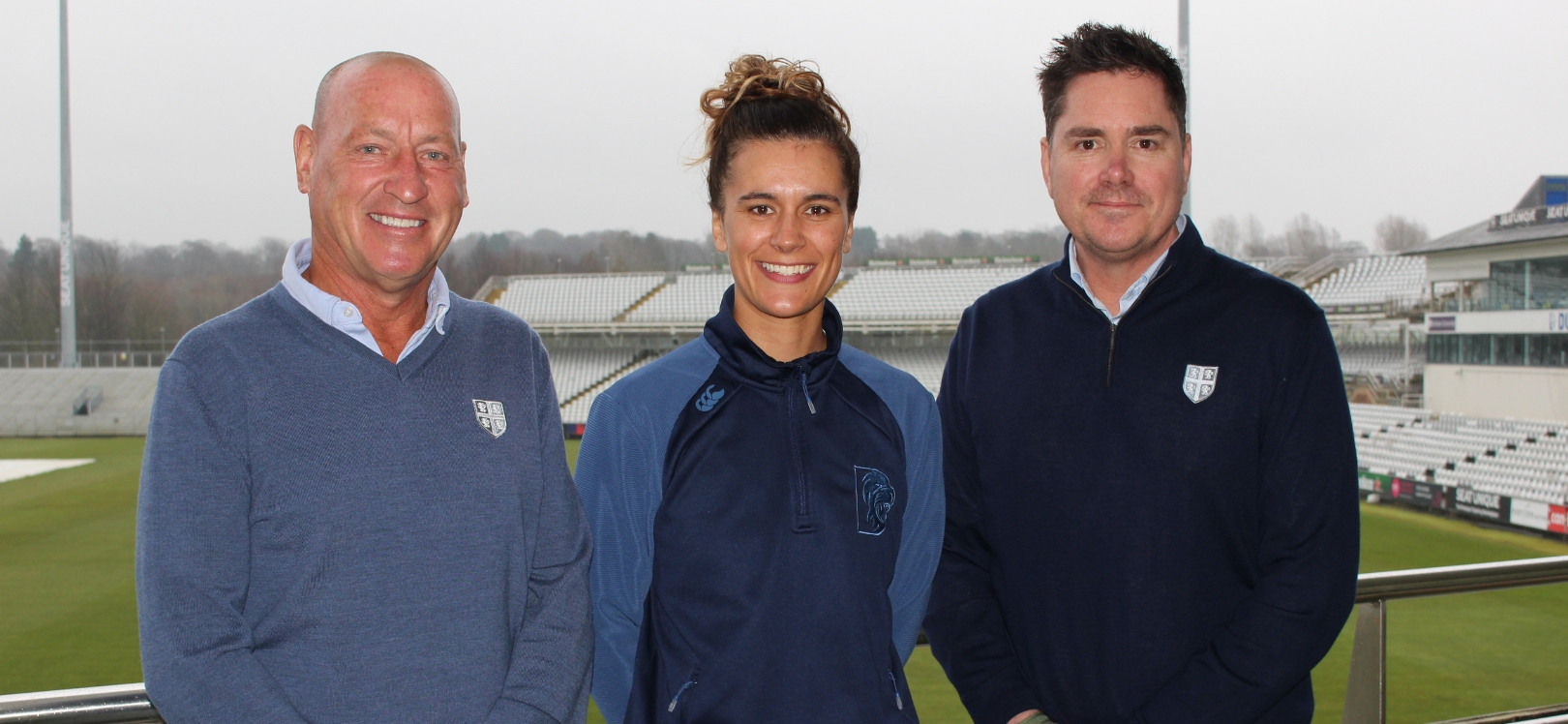 Three people stood in front of a cricket ground